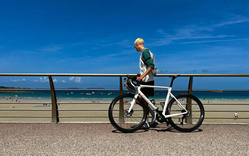 ein junger, blonder Mann, der ein Rennrad am Geländer zum Strand entlangschiebt