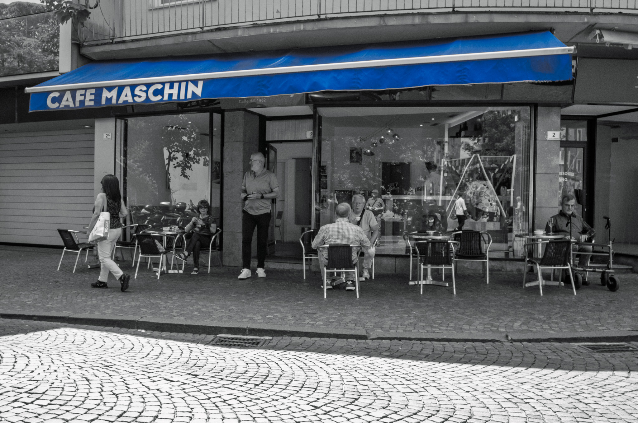 ein Straßenkaffee, auf dessen Sonnenmarkise “Cafe Maschin” steht, davor Leute sitzend