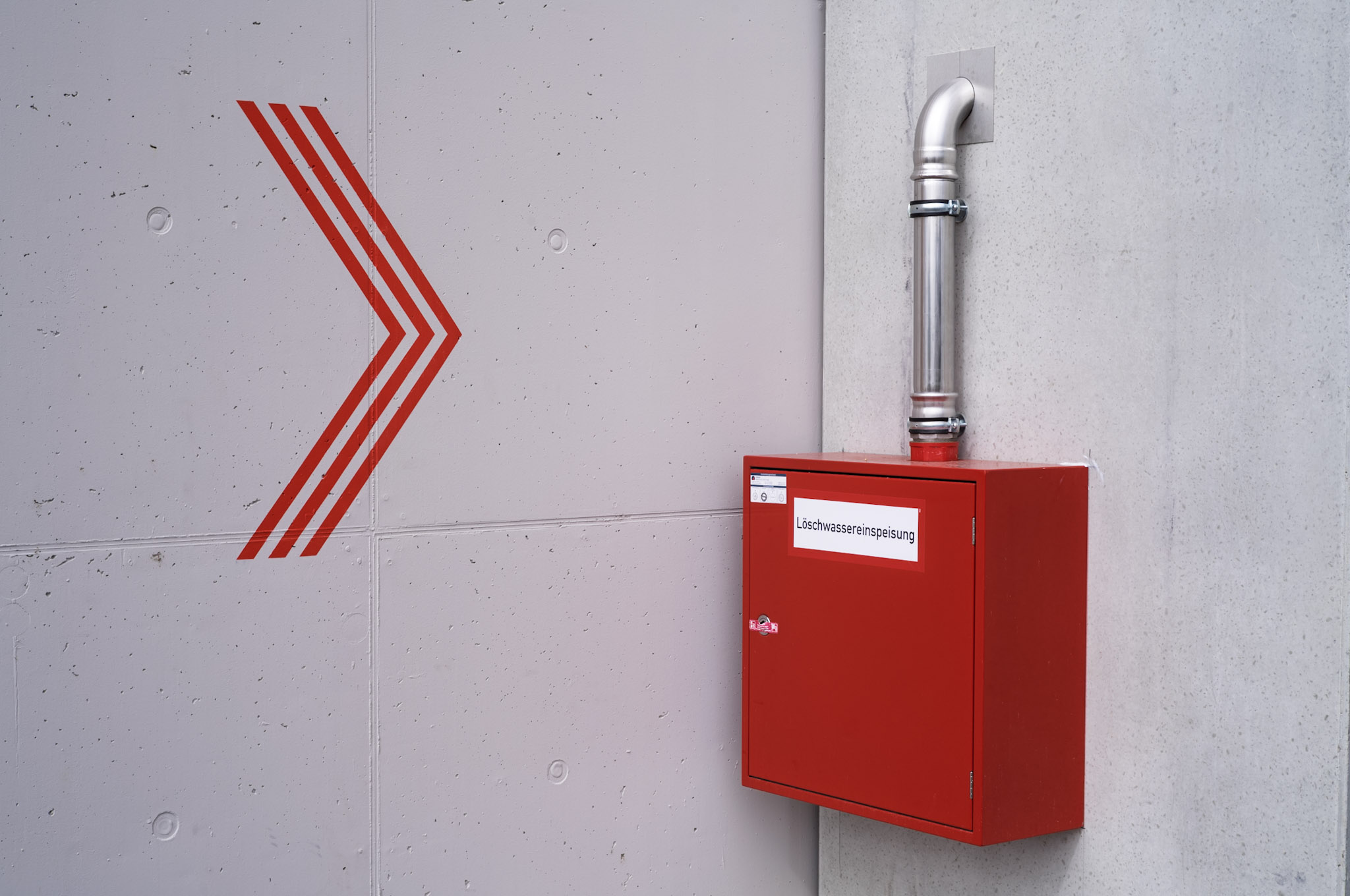 ein roter Metallkasten mit der Aufschrift “Löschwassereinspeisung” an einer makellos sauberen, neuen Betonwand, links daneben ein großer roter nach rechts weisender Pfeil