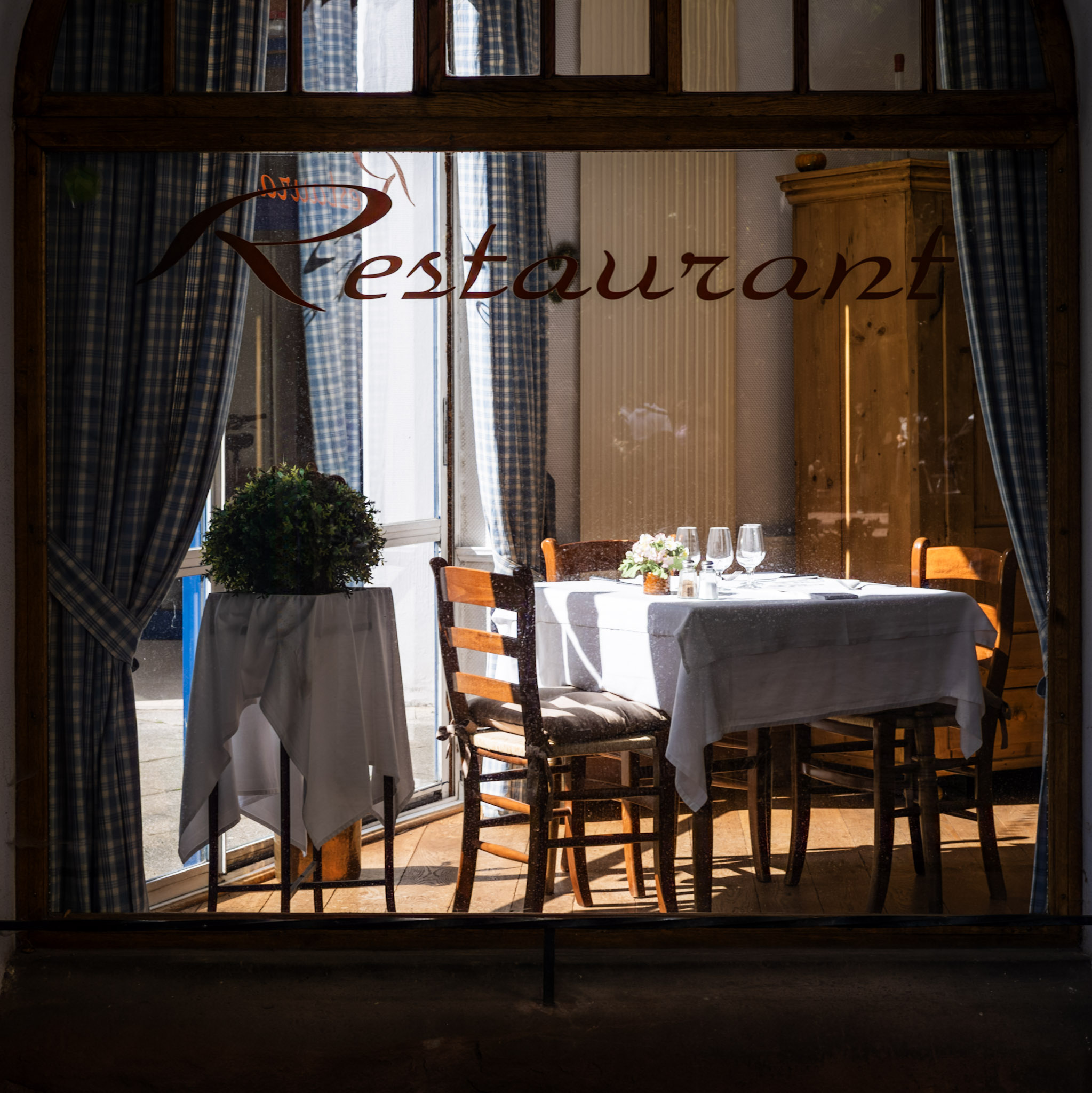 iBlick durch eine Fensterscheibe auf einen kleinen quadratischen Tisch, eingedeckt für 3 Personen, an der Wand eine karierte Gardine. iAuf der Fensterscheibe steht in großen Buchstaben “Restaurant”.