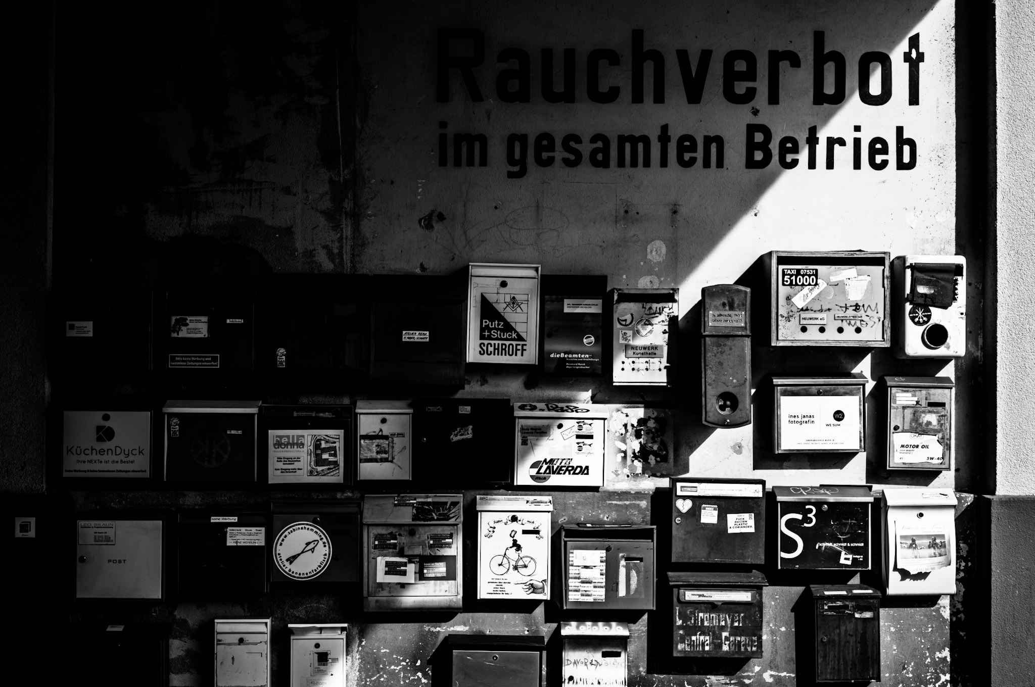 Viele Firmenbriefkästen an einer Wand, auf die ein harter Schlagschatten fällt, so dass eine diagonale Linie Licht und Schatten trennt. Auf der Wand steht “Rauchverbot im gesamten Betrieb” geschrieben.